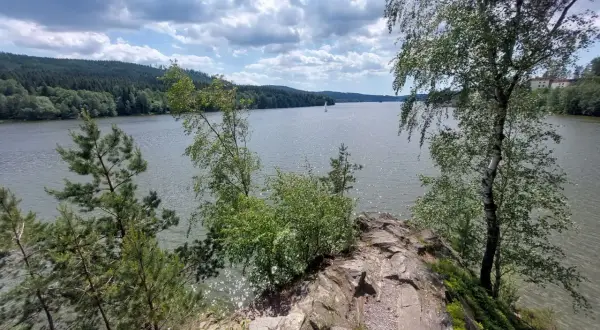 Felsiger Uferrand am Moldau Stausee mit Bäumen und Segelbooten auf dem glitzernden Wasser unter bewölktem Himmel.