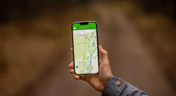 Dłoń trzyma smartfon z mapą i trasą. Aplikacja do planowania podróży pokazuje ścieżkę przez tereny zielone i wodne.