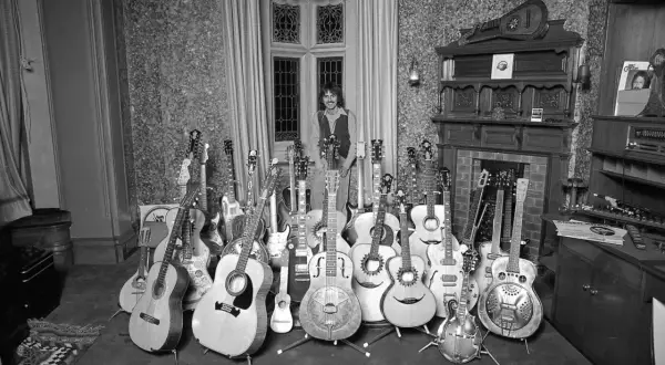 Mann mit vielen Rock n Roll Instrumenten, darunter Akustik- und E-Gitarren, vor einem Fenster.