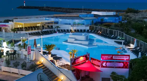Kvällsbild över en poolklubb vid Cap d'Agde. Ljusblå pool med solstolar, palmer och en scen i mitten. Havet och en fyr syns i bakgrunden.