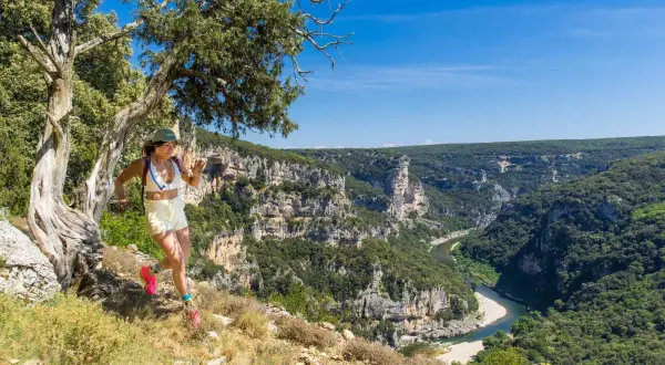 Une femme court sur un sentier escarpé avec une vue spectaculaire sur le **trail gorge de l'Ardèche**. Le soleil brille sur les falaises et la rivière.