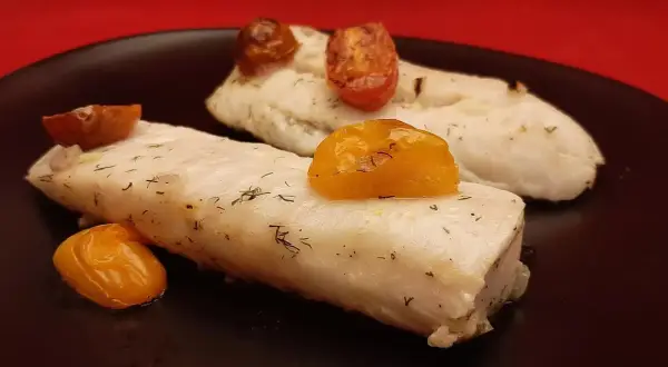 Deux pavés de dos de merlu tendres, parsemés d'aneth et surmontés de tomates cerises rôties, sur une assiette noire.
