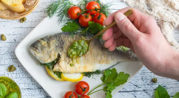 Eine goldbraune Dorade in der Pfanne, garniert mit frischen Kräutern, Kirschtomaten und Zitronenscheiben. Eine Löffelspitze grüner Sauce wird auf den Fisch gegeben.