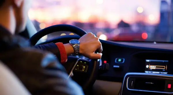 Förare med klocka styr bil med automatlåda genom stadens ljus i skymningen.