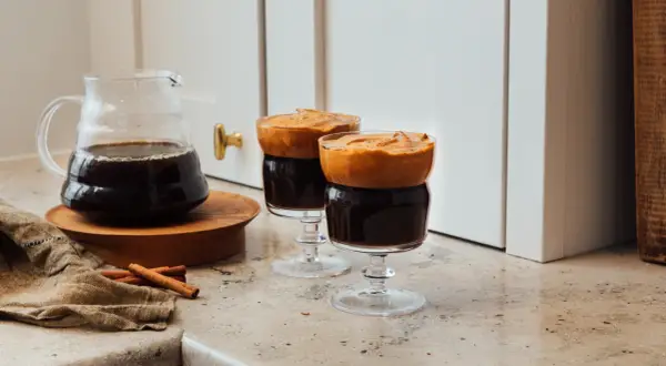 Två glas med mörkt, kallt kaffe toppat med fluffigt skum. En karaff med kaffe och kanelstänger står bredvid.