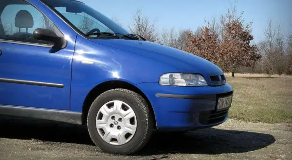 Błękitny Fiat Albea stoi na polnej drodze, w tle jesienne drzewa.