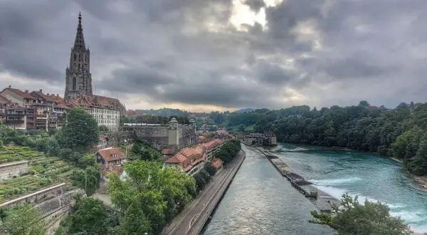 Blick von der Besucherplattform über Bern: Münster, Aare und grüne Hänge unter dramatischem Himmel.