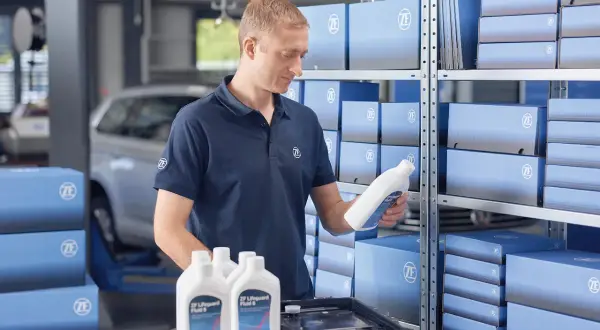 Mekaniker håller en flaska ZF Lifeguard Fluid 8, perfekt för att byta olja i automatlåda. Hyllor fyllda med ZF-produkter.