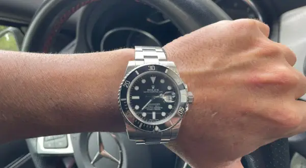 Mano con reloj Rolex Submariner, símbolo de invertir en relojes de lujo. El volante de un coche de alta gama sugiere un estilo de vida exitoso.