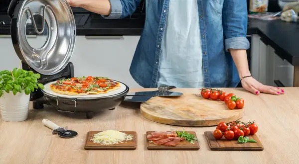 Osoba przygotowuje pizzę w piecyku, obok składniki: pomidorki, ser, szynka.
