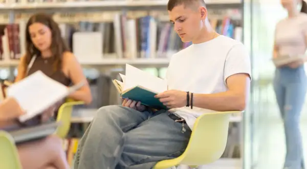 Młody mężczyzna w białej koszulce i dżinsach czyta książkę w bibliotece. To obraz jak przedwiośnie, jaka epoka – czas nauki i rozwoju.