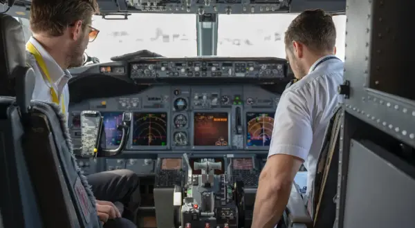 Dwóch pilotów samolotu w kokpicie. Jeden z nich, pilot, patrzy na instrumenty, drugi obsługuje sterowanie.