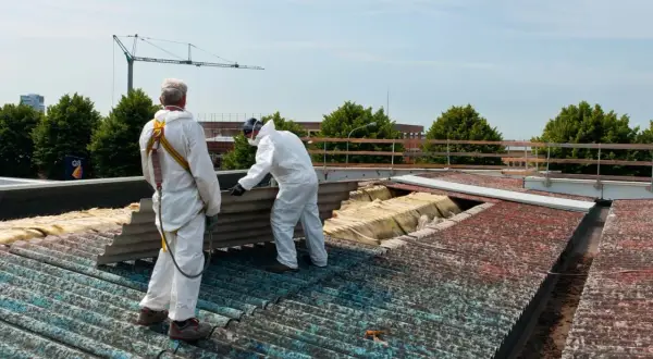 Dwóch pracowników w kombinezonach ochronnych usuwa eternit z dachu. Kiedyś popularny materiał budowlany, dziś wymaga specjalistycznej utylizacji.