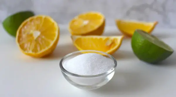 Ciotolina di acido citrico cristallino circondata da spicchi di limone e lime, ingredienti freschi per un tocco frizzante.