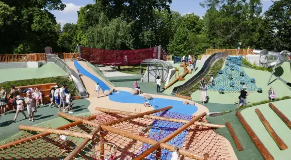 Kolorowy plac zabaw na Alejach Ujazdowskich z licznymi zjeżdżalniami, siatkami do wspinaczki i wodnymi elementami, gdzie bawią się dzieci.