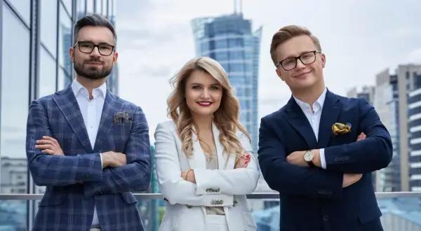 Trzy osoby w eleganckich garniturach stoją na tle nowoczesnych budynków.