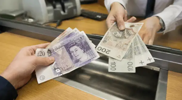 Ręka podaje wycofane funty i polskie banknoty w okienku bankowym.