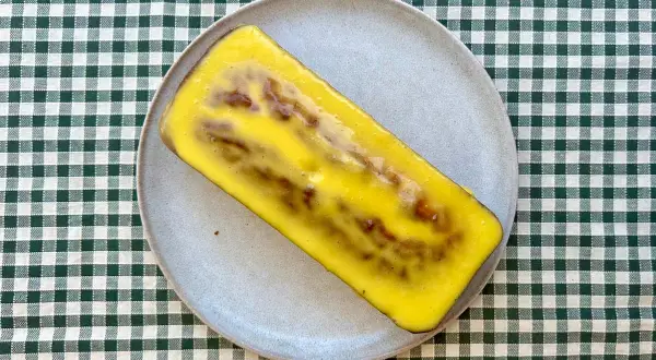 Un delicioso bizcocho de mango y yogur, cubierto con un glaseado amarillo brillante, descansa sobre un plato gris.