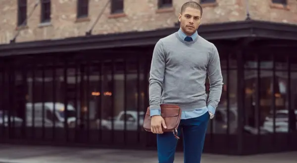 Ein Mann mit Stil, der sein **Outfit zusammenstellen Herren** perfektioniert: grauer Pullover, hellblaues Hemd, blaue Hose und eine braune Ledertasche.