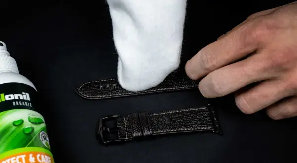 Ein weißes Tuch poliert ein dunkles Lederarmband. Daneben steht eine Flasche "Collonil Organic Protect & Care" zur Pflege.