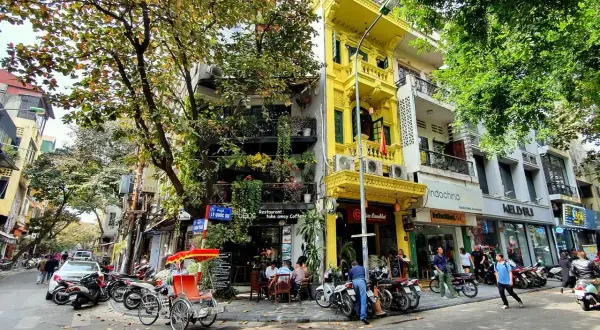 Ulica w Hanoi z żółtą kamienicą, rikszą i skuterami. W tle widać sklepy i restauracje, gdzie można wymienić walutę w Hanoi.