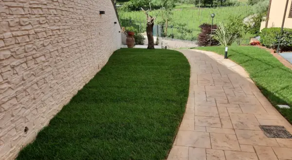 Sentiero in cemento stampato spessore minimo, con finitura effetto pietra, fiancheggiato da prato verde e muro in pietra grezza.