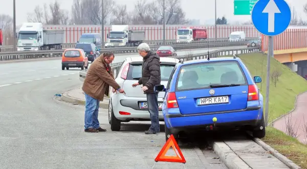 Dwóch mężczyzn dyskutuje przy uszkodzonych samochodach po kolizji. Trójkąt ostrzegawczy sugeruje, że może być spowodowanie kolizji mandat.