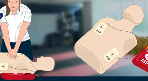 Ilustracja przedstawia manekina do resuscytacji z przyklejonymi elektrodami AED oraz czerwony defibrylator AED.