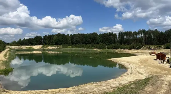 Zbiornik Czarna Sędziszowska z piaszczystą plażą i lasem w tle. Błękitne niebo z białymi chmurami odbija się w spokojnej tafli wody.