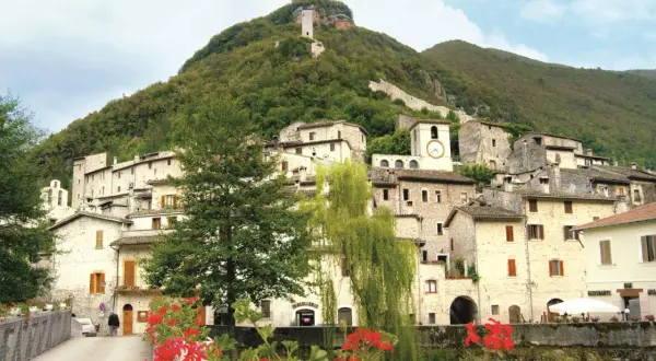 Antico borgo arroccato su un colle verdeggiante, uno dei paesi della Valnerina, con case in pietra, un campanile con orologio e una torre medievale in cima.