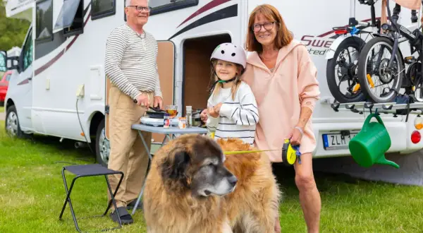 Familjen njuter av att campa med hund vid husbilen. En flicka med hjälm, en man som lagar mat och en kvinna med koppel till hunden.