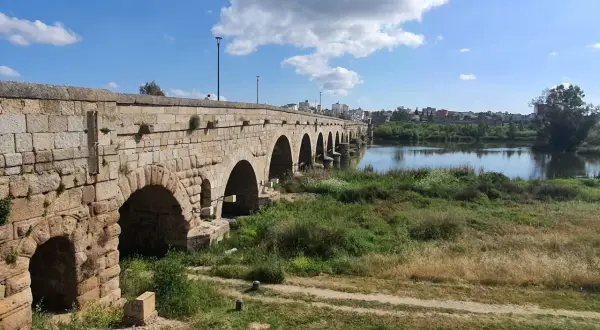 Puente romano de piedra con arcos sobre un río, bordeado de vegetación. Un camino de tierra serpentea por la orilla, evocando la antigua **Vía de la Plata**.