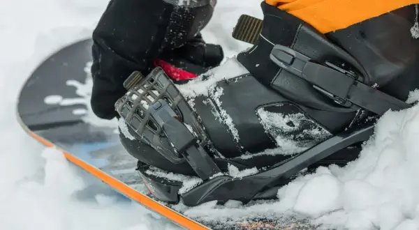 Rękawica zapina but snowboardowy na desce, przygotowując się do wygrzewania butów snowboardowych w śniegu.