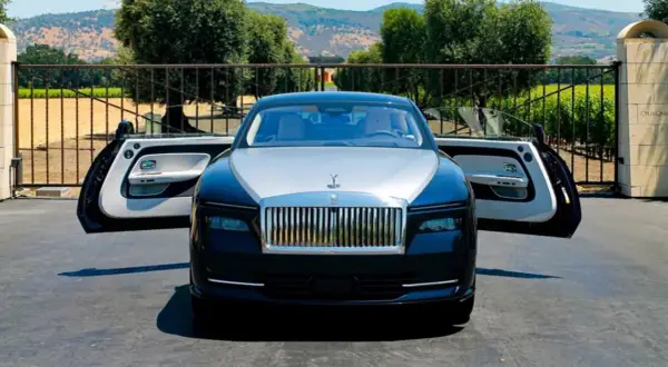 Elegancki Rolls-Royce Spectre z otwartymi drzwiami, zaparkowany przed bramą posiadłości z widokiem na wzgórza.