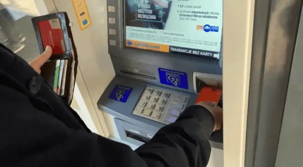Osoba wkłada kartę do bankomatu, by wypłacić pieniądze. Widoczny portfel z kartami.