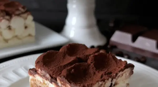 En bit krämig tiramisu, perfekt för ett tiramisu recept original. Pudrad med kakao, med kaffebönor och choklad i bakgrunden.