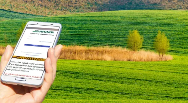 Dłoń trzyma smartfon z aplikacją ARiMR do składania wniosków o płatności bezpośrednie na tle zielonych pól.