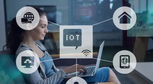 Kobieta pracuje przy laptopie, otoczona ikonami symbolizującymi internet rzeczy (IoT): dom, chmura, ciężarówka, smartfon.