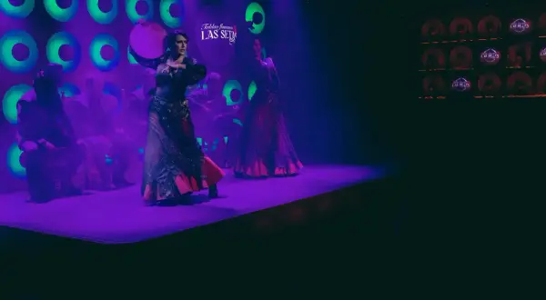Dos bailarinas de flamenco, una con un tambor, aprenden a bailar sevillanas en un escenario con luces moradas y verdes.