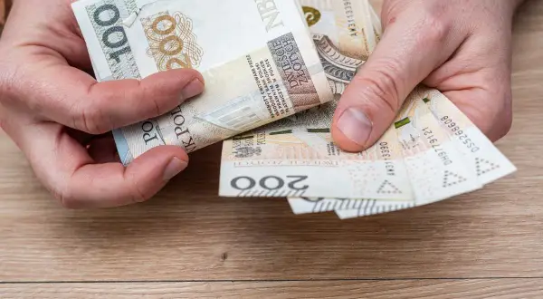 Dłonie trzymają polskie banknoty 100 i 200 zł. Może to być pomoc w opłaceniu rachunków, np. bon ciepłowniczy.