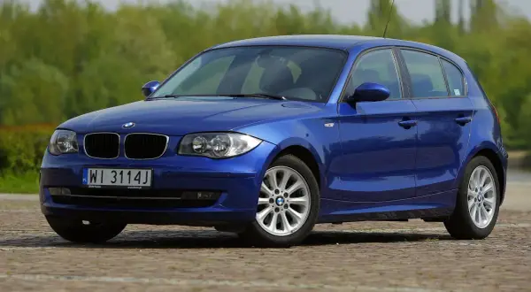Niebieskie BMW E87 z polskimi tablicami WI 3114J, z nowymi żarówkami w reflektorach, zaparkowane na bruku.
