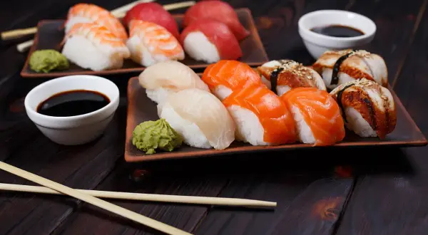 Jak wygląda sushi: na ciemnym drewnie ułożone są talerze z nigiri: krewetka, łosoś, biała ryba i węgorz. Obok sos sojowy i wasabi.