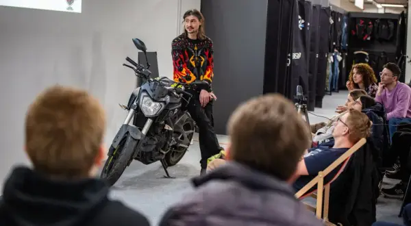 Mężczyzna w bluzie z płomieniami siedzi na motocyklu przed publicznością.