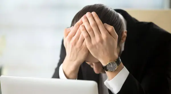 Pracownik w garniturze z rękami na twarzy, przy laptopie. Wygląda na przygnębionego, być może z powodu odpowiedzialności finansowej pracownika za błędy.