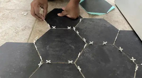 Hands place dark hexagonal tiles over existing floor tiles, using white spacers to ensure even gaps.