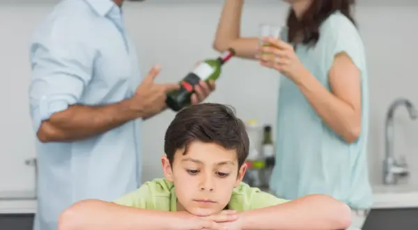 Smutny chłopiec opiera głowę na rękach, podczas gdy rodzice w tle piją alkohol. Obraz symbolizuje trudne dzieciństwo i problemy związane z dda.