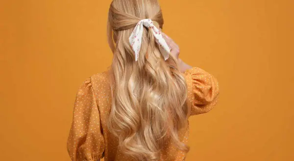 Chevelure blonde ondulée, coiffée en demi-queue avec un foulard fleuri, sur fond orange.