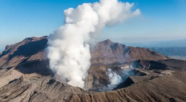 Fumée s'échappant du cratère du volcan Aso, sous un ciel bleu clair avec la lune visible.