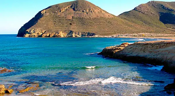 Aguas turquesas bañan la costa rocosa de el playazo las negras, con montañas áridas de fondo bajo un cielo azul.