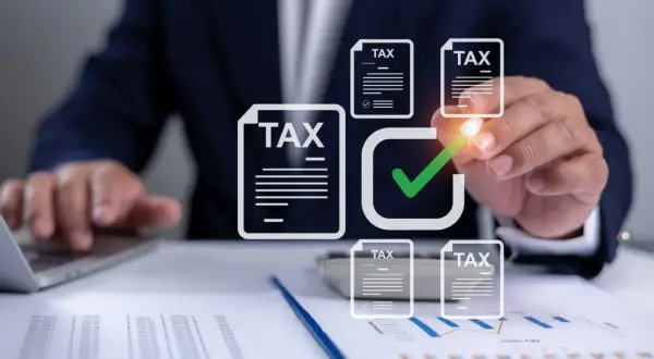 Dłoń zaznacza zielonym znacznikiem dokumenty z napisem "TAX" na tle wykresów i laptopa.